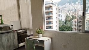 A small kitchen with a stove, microwave, and a dining table with a chair. A window offers a view of a cityscape with buildings and trees. - Apartment in Jounieh, sea view, 6th floor, wifi (Keserouan)