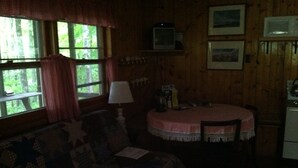 A cozy room with a wooden interior, a round table with a checkered tablecloth, a sofa with a quilt, and a window with curtains. - Secluded Cabin Nestled In The Trees Overlooking Fishing & Canoe Pond (Burlington Flats)