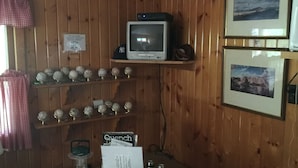A room with wooden walls, a television on a shelf, baseballs on display, a table with various items, and framed pictures on the wall. - Secluded Cabin Nestled In The Trees Overlooking Fishing & Canoe Pond (Burlington Flats)
