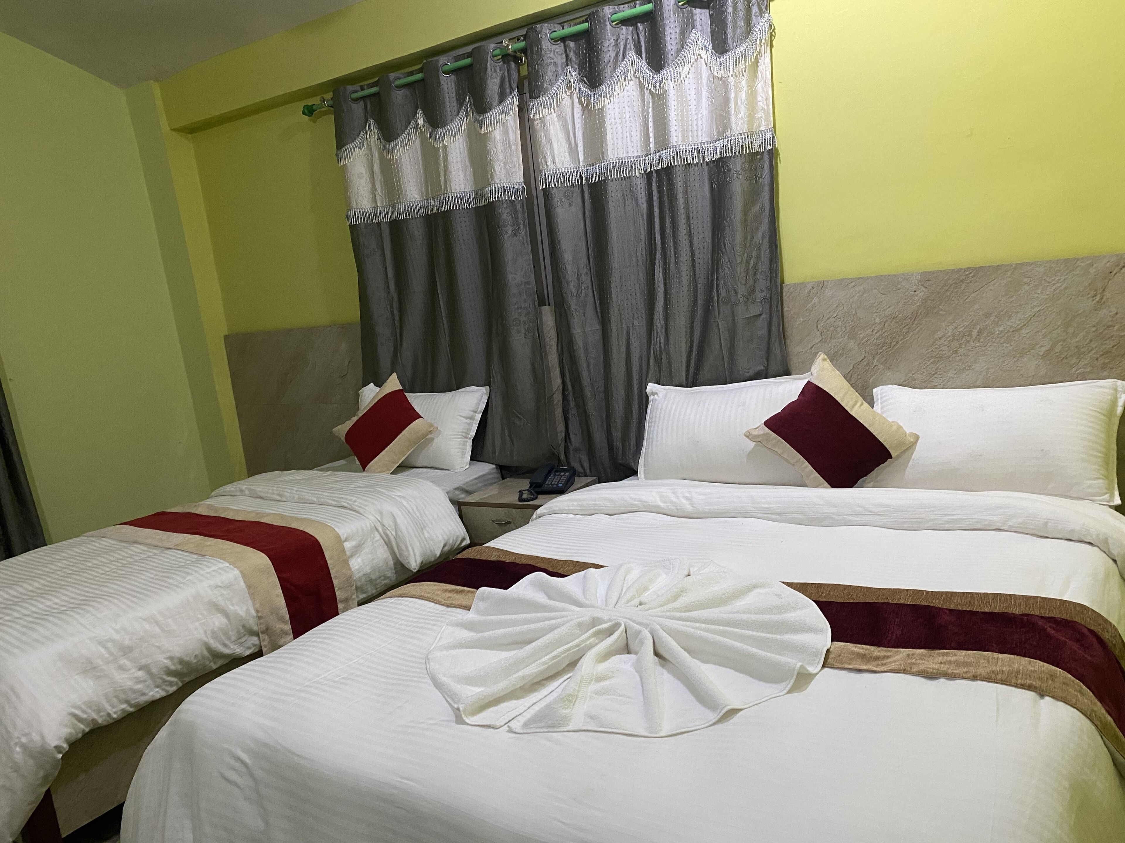 Habitación de hotel con dos camas, cada una con ropa de cama blanca y almohadas rojas y beige. Hay una cortina verde con un adorno de borla, y una toalla blanca sobre la cama en primer plano.