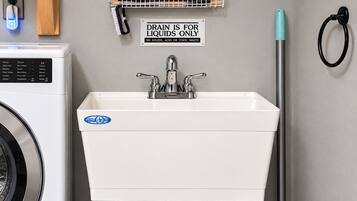 A laundry room with a sink, a sign indicating the drain is for liquids only, a mop, and a broom.