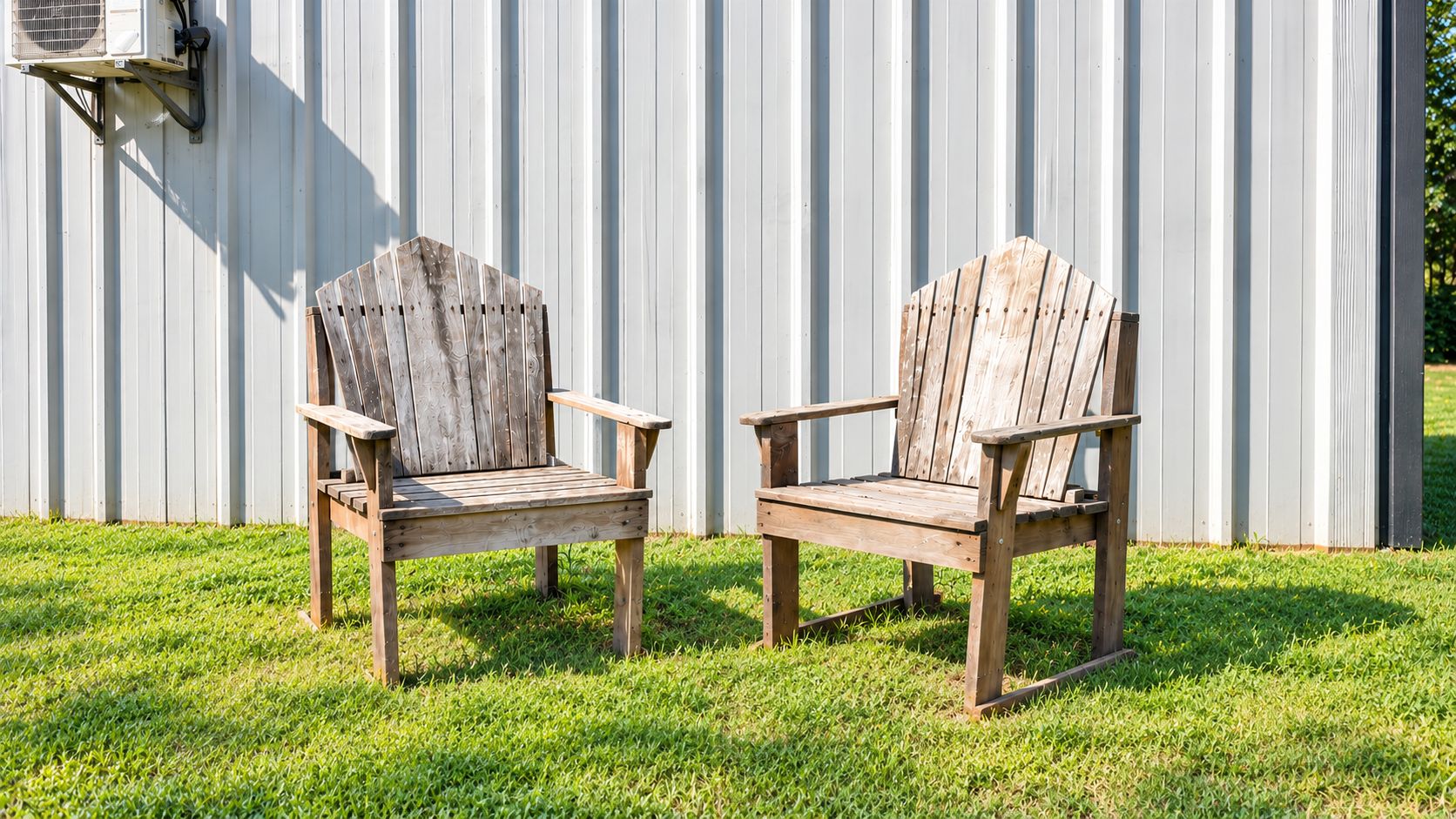 Two wooden Adirondack chairs with metal frames, positioned on a grassy lawn in front of a corrugated metal wall.