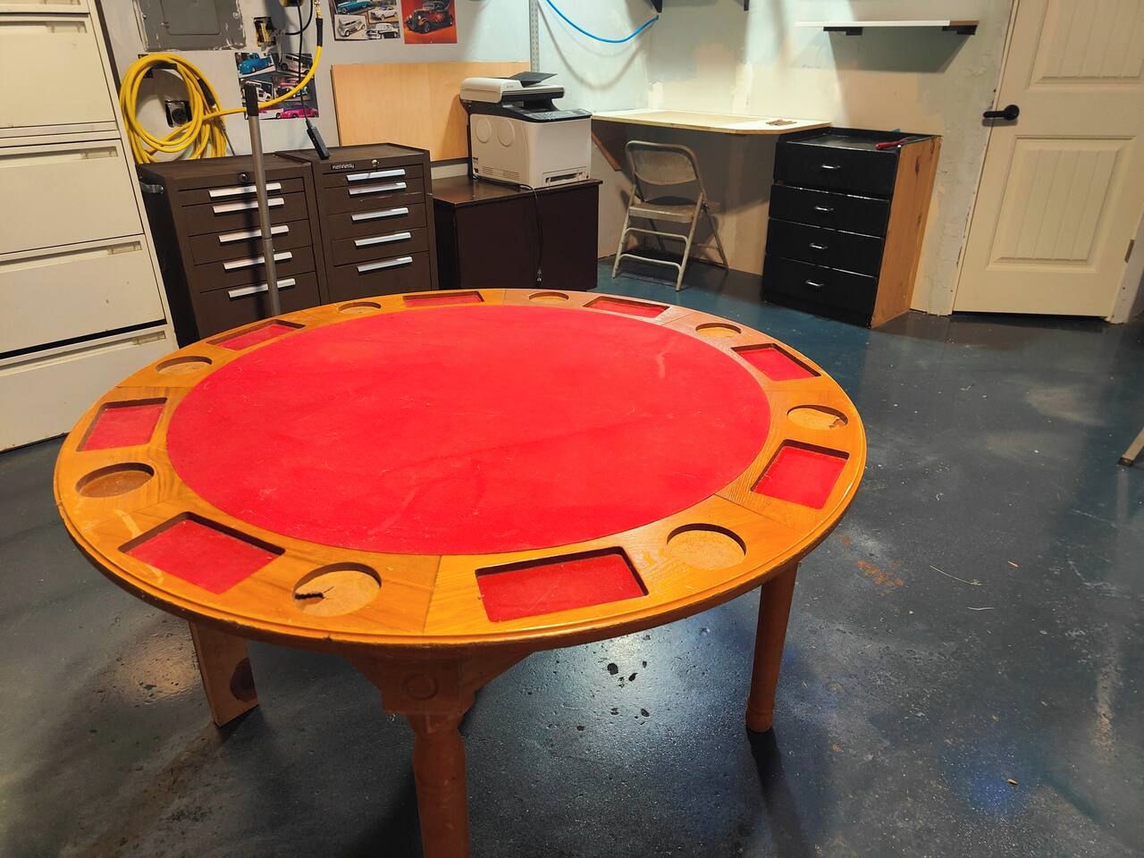 A round table with a red top and wooden edges, surrounded by drawers and a printer in a workshop-like setting.