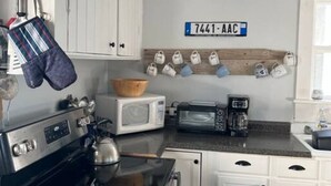 A kitchen with white cabinets, a microwave, a toaster, and a coffee maker. There is a wooden shelf with shoes and a sign with the number 7441-AAC. - The Webster House Vacation Station (Vinalhaven)