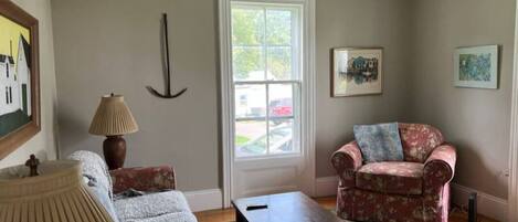 A living room with a sofa, armchair, coffee table, lamp, and paintings on the walls.