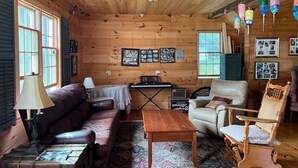 A cozy cabin interior with a wooden ceiling, a leather sofa, a white armchair, a small table, and a keyboard. - The Webster House Vacation Station (Vinalhaven)