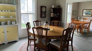 A dining room with a wooden table, six chairs, a clock, a bookshelf, and a window. - The Webster House Vacation Station (Vinalhaven)