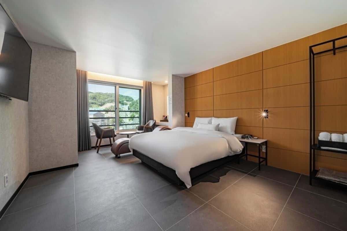 Ein modernes Hotelzimmer mit einem großen Bett, einem Schreibtisch, einem Sessel und Blick ins Freie.