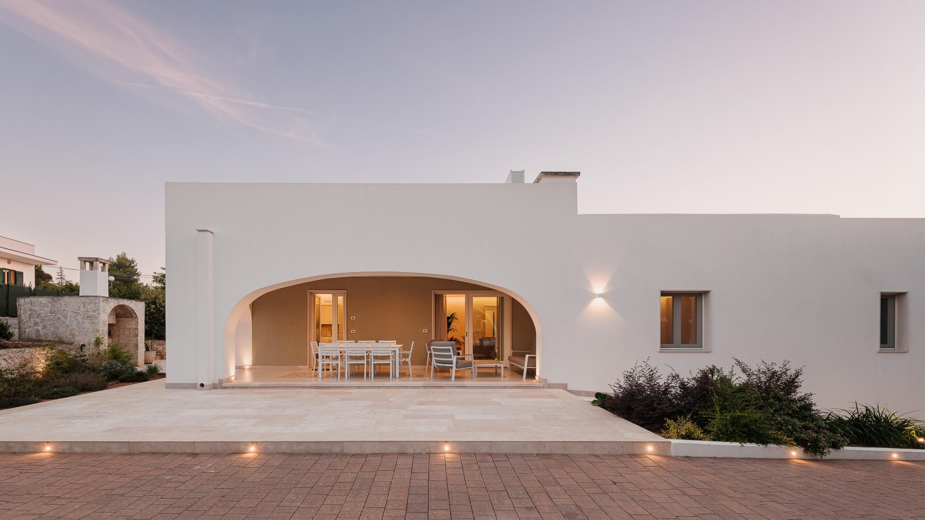 Une maison moderne dotée d’une entrée en arc de cercle, d’un patio avec mobilier d’extérieur et d’un intérieur bien éclairé.