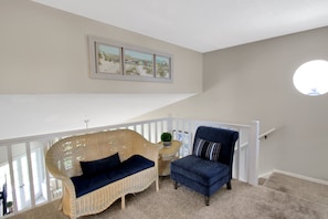 Una habitación con un sofá de mimbre, un sillón azul, una mesita con una planta y una escalera con barandilla blanca.