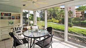 A sunlit room with a glass table and chairs, a ceiling fan, a bookshelf, and a view of a well-maintained garden through large windows. - Relaxing Siesta Key condo - pickleball - 2 heated pools - beach access - GBB 1344 (Siesta Key)