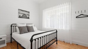 Ein Schlafzimmer mit Metallbettgestell, weißer Bettwäsche, einem karierten Kissen, einem Nachttisch, einem Fenster mit durchsichtigen Vorhängen und einem Wandhaken für Kleidung.