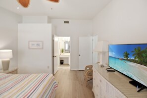 Habitación de hotel con cama, televisor mostrando una escena de playa, una silla pequeña y un baño visible a través de una puerta abierta.