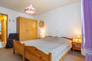 A bedroom with a wooden bed, a chair, a wardrobe, a nightstand with a lamp, and a ceiling light fixture.