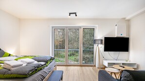 Una habitación de hotel moderna con cama, sofá, televisor y balcón con vistas a árboles.