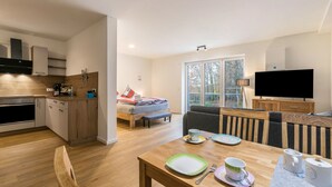 Une cuisine moderne avec des meubles en bois, un coin repas avec une table dressée pour deux personnes, un canapé et un téléviseur dans un salon doté d’une grande fenêtre.