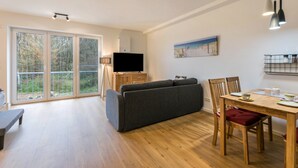 Un salon moderne comprenant un canapé, une table à manger et des chaises, un téléviseur à écran plat et de grandes fenêtres donnant sur la verdure.