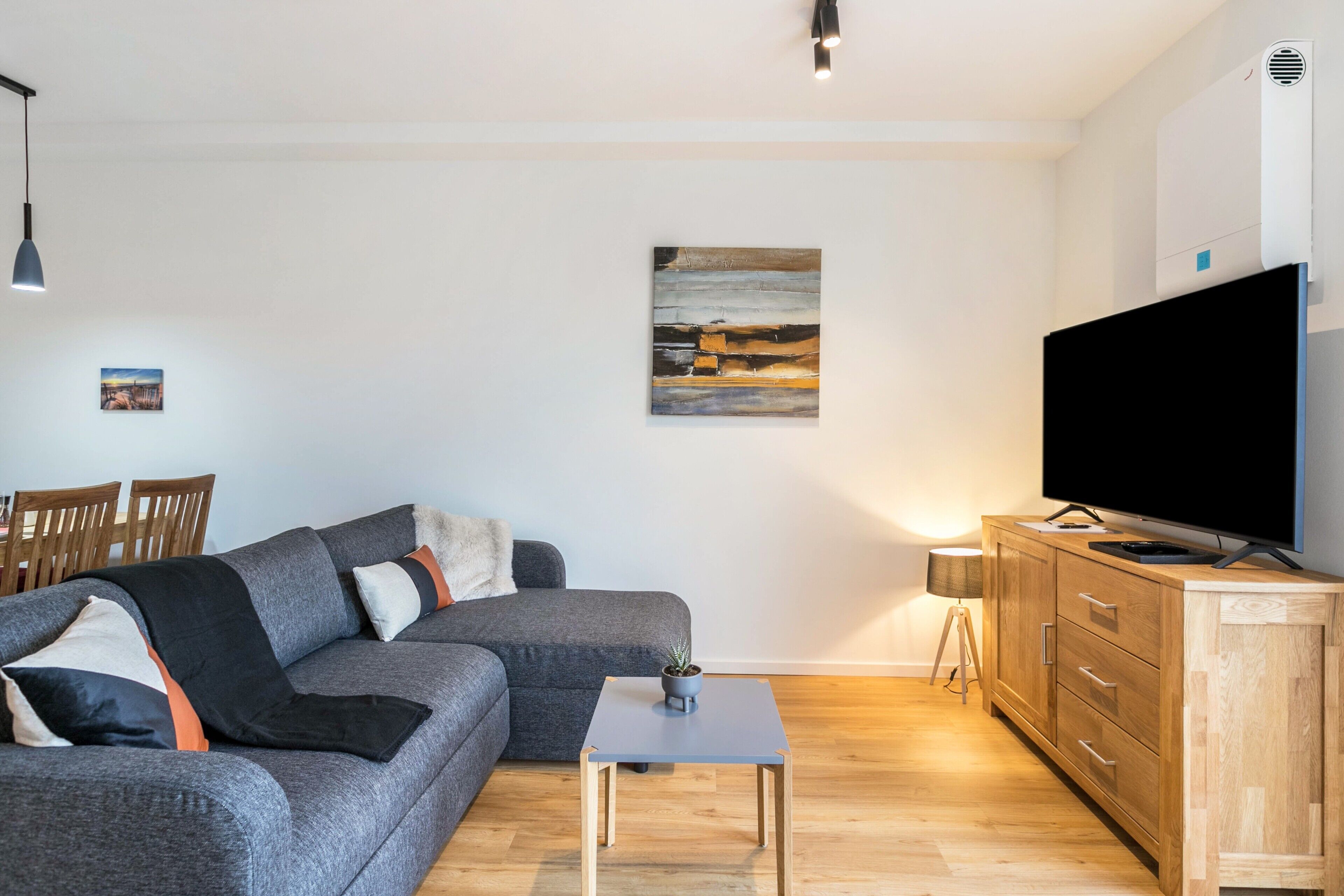Una sala de estar moderna con un sofá gris, un mueble de madera, un televisor de pantalla plana y una mesita con una planta.