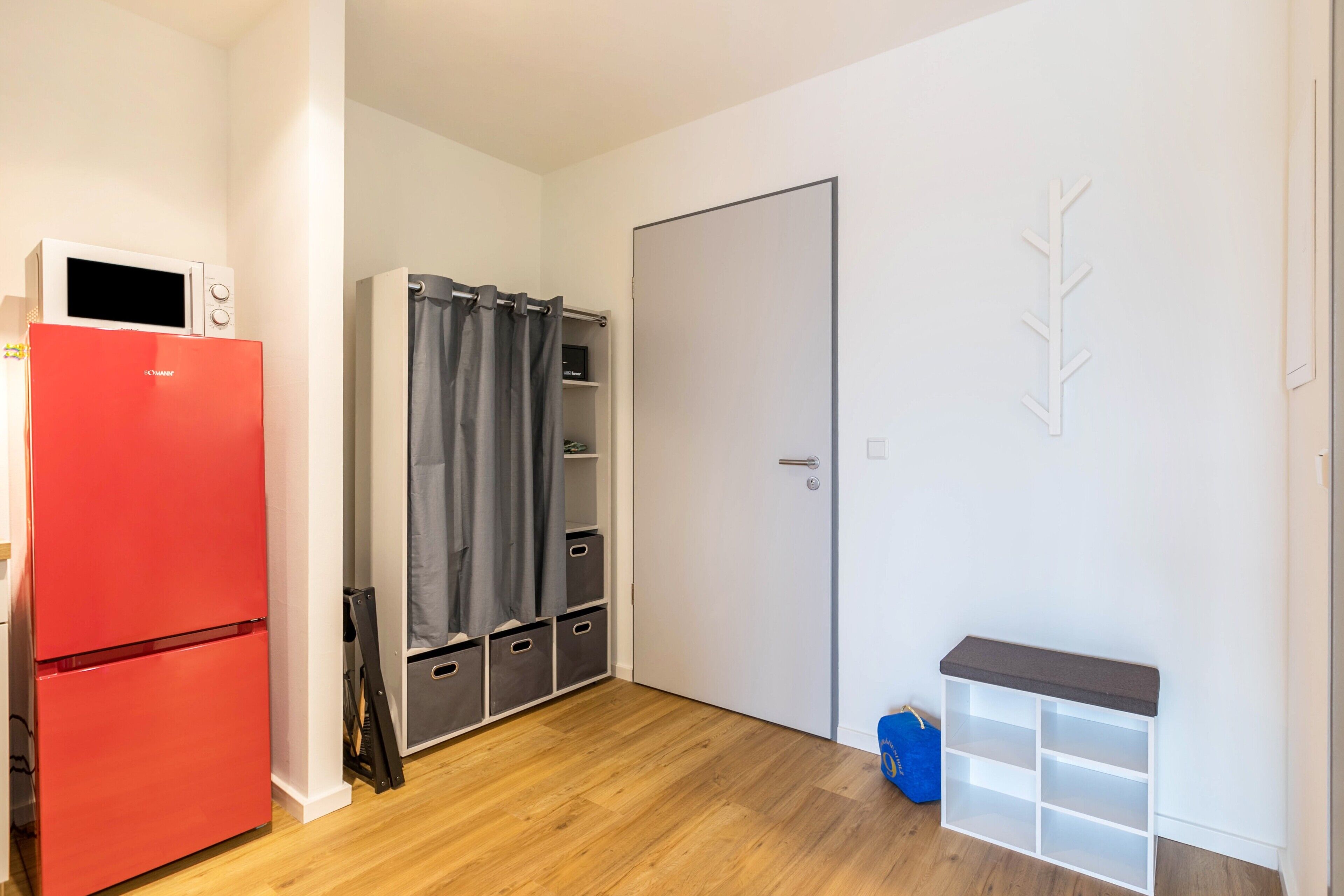 Una habitación compacta con un refrigerador rojo, un armario gris, una puerta blanca y una pequeña estantería blanca.