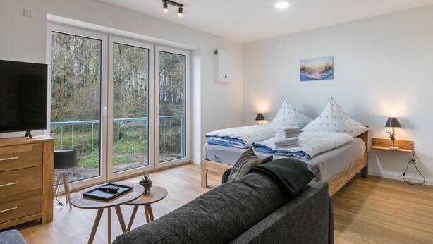 Un dormitorio moderno con cama, lámparas de noche, televisión, sofá y un balcón con vistas a árboles.