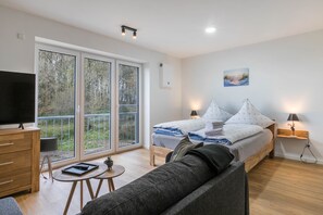 Een moderne slaapkamer met een bed, nachtlampjes, een televisie, een bank en een balkon met uitzicht op bomen.