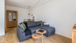 Una sala moderna con un sofá seccional gris, una mesita redonda de madera y un comedor al fondo.