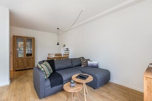 A modern living room with a grey sectional sofa, a small round wooden table, and a dining area in the background.