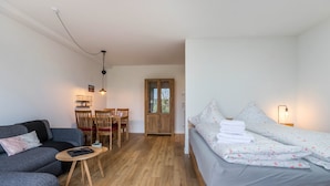 Habitación compacta con sofá gris, una pequeña mesa de madera, cama con toallas ordenadas, armario de madera y zona de comedor con sillas.