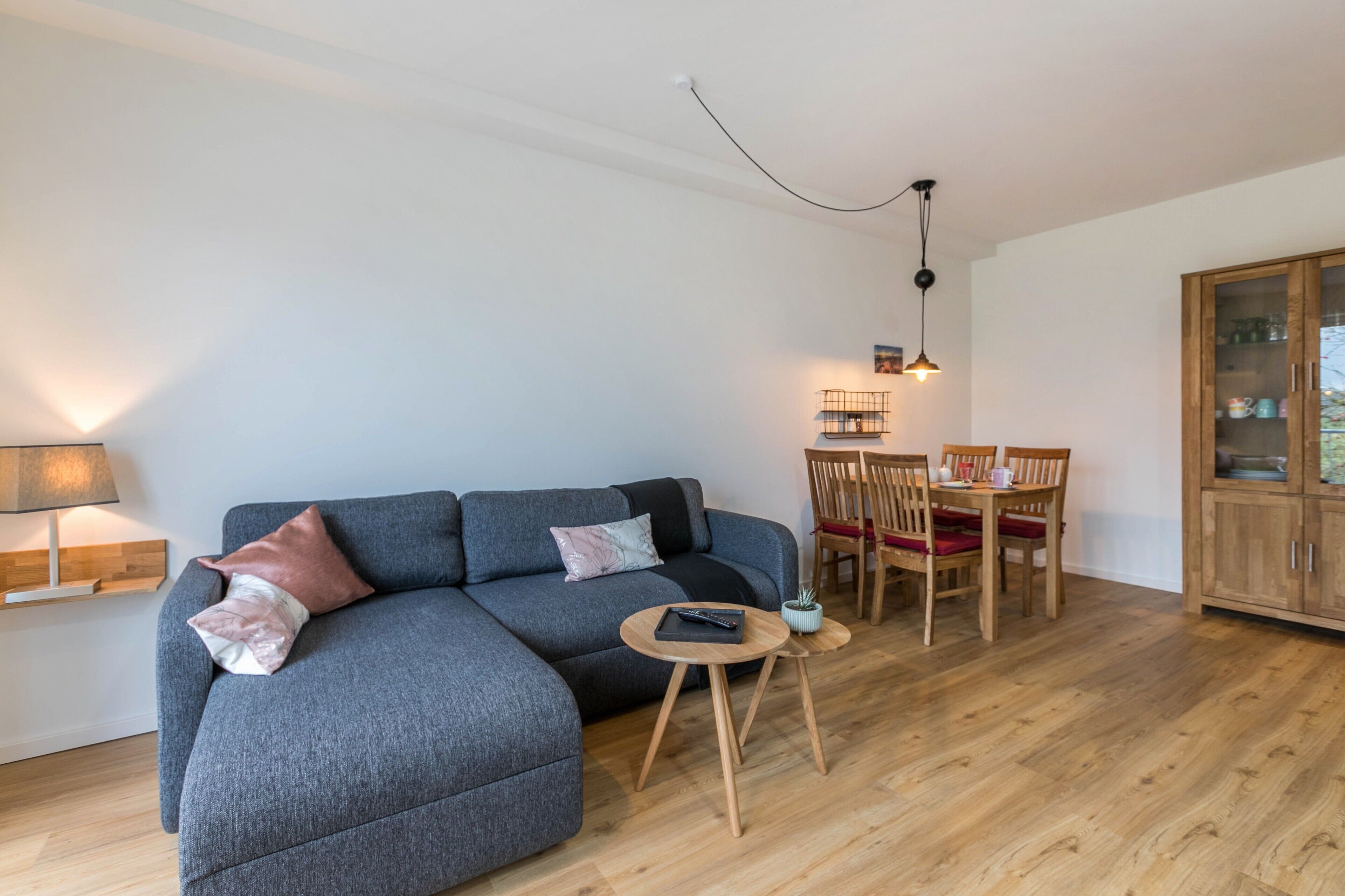 A modern living room with a grey sectional sofa, a small round wooden table, a dining table with chairs, and a cabinet with glass doors.