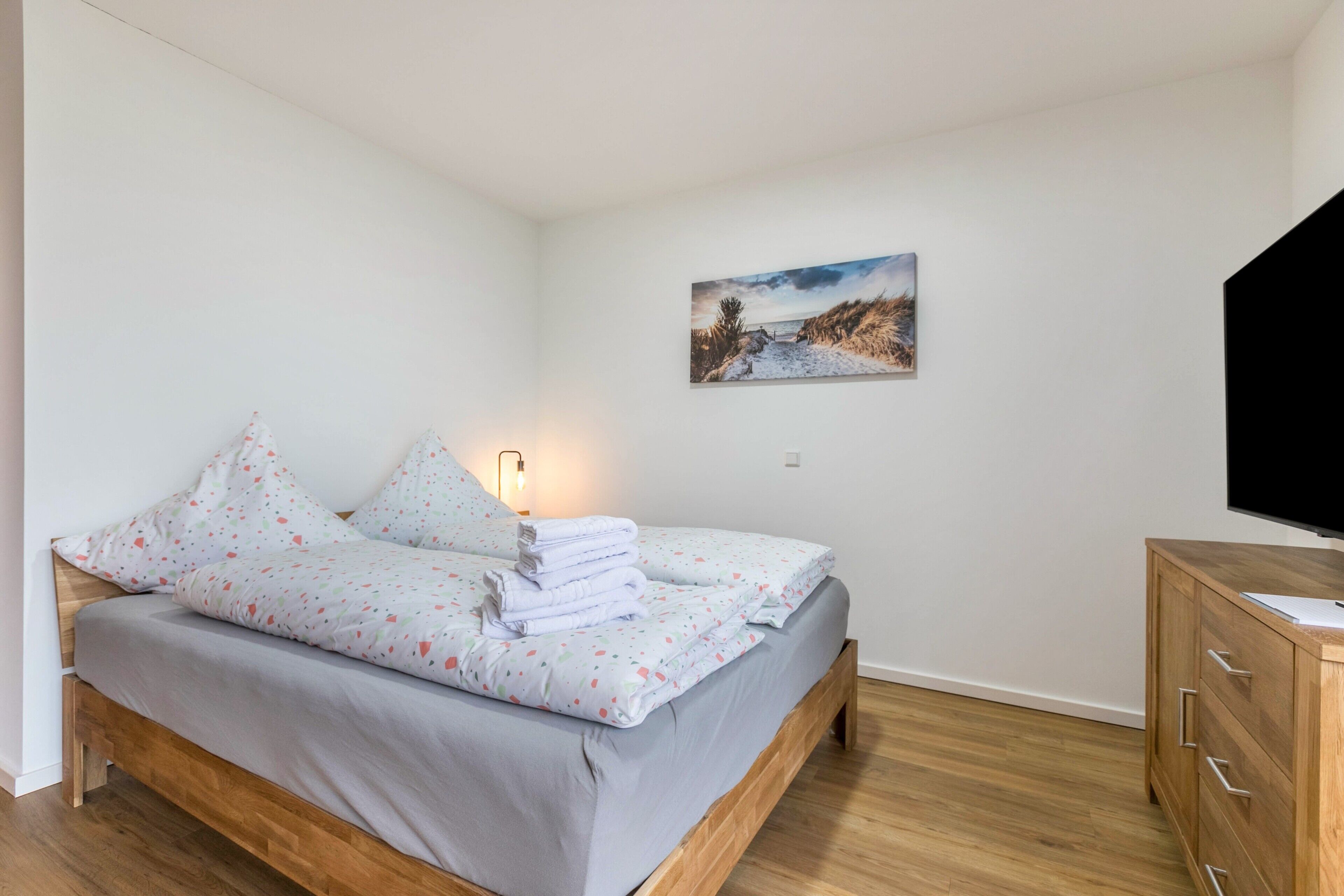 A bedroom with a bed, a wooden dresser, a painting on the wall, and a television.