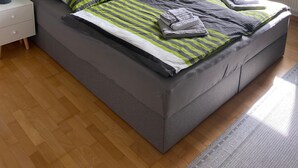 Ein Schlafzimmer mit einem grau gepolsterten Bett, gestreifter Bettwäsche, einem Nachttisch mit Lampe, einem Fenster mit Jalousien und einer Pendelleuchte.