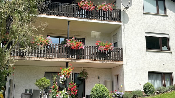 Ein zweistöckiges Gebäude mit Balkonen, die mit Blumen geschmückt sind, einer Satellitenschüssel und einem Gartenbereich mit Kübelpflanzen.