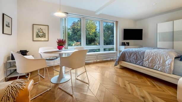 Un dormitorio moderno con cama, televisor, zona de comedor con sillas y un ventanal grande.