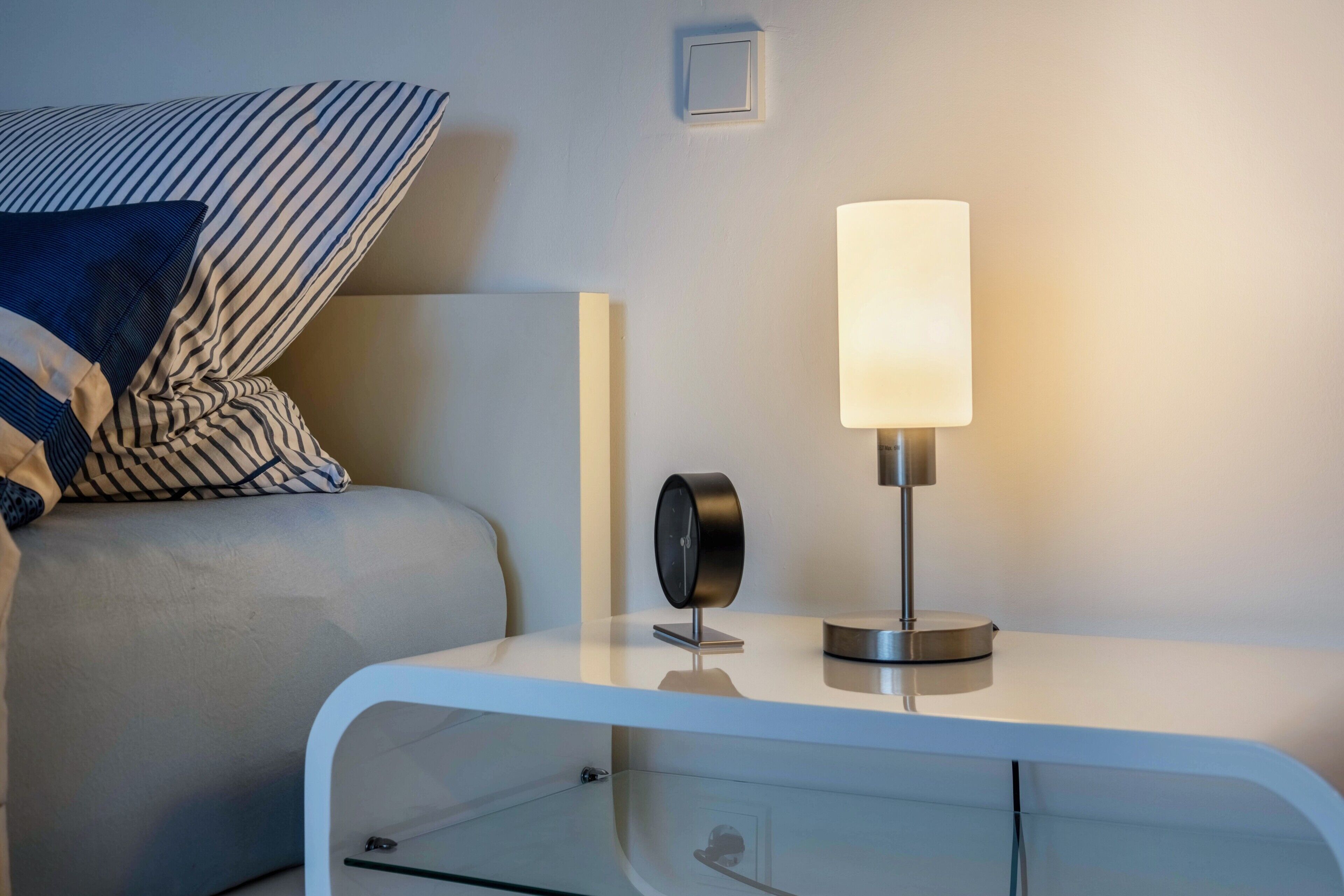 Une table de chevet moderne avec une lampe, une horloge et un oreiller sur un lit.