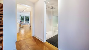 Un interior moderno con suelo de madera, ducha de vidrio y una zona de cocina visible a través de un umbral abierto.