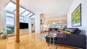 Ein geräumiger Wohnbereich mit großem Fenster, einer dunklen Couch, einem runden Couchtisch und einer Küche im Hintergrund.
