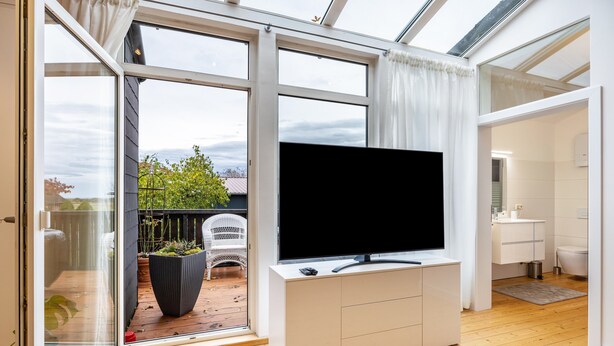Ein modernes Zimmer mit einem großen Fernseher auf einem weißen Schrank, einer Schiebetür zu einem Außenbereich und einem sichtbaren Badezimmer.