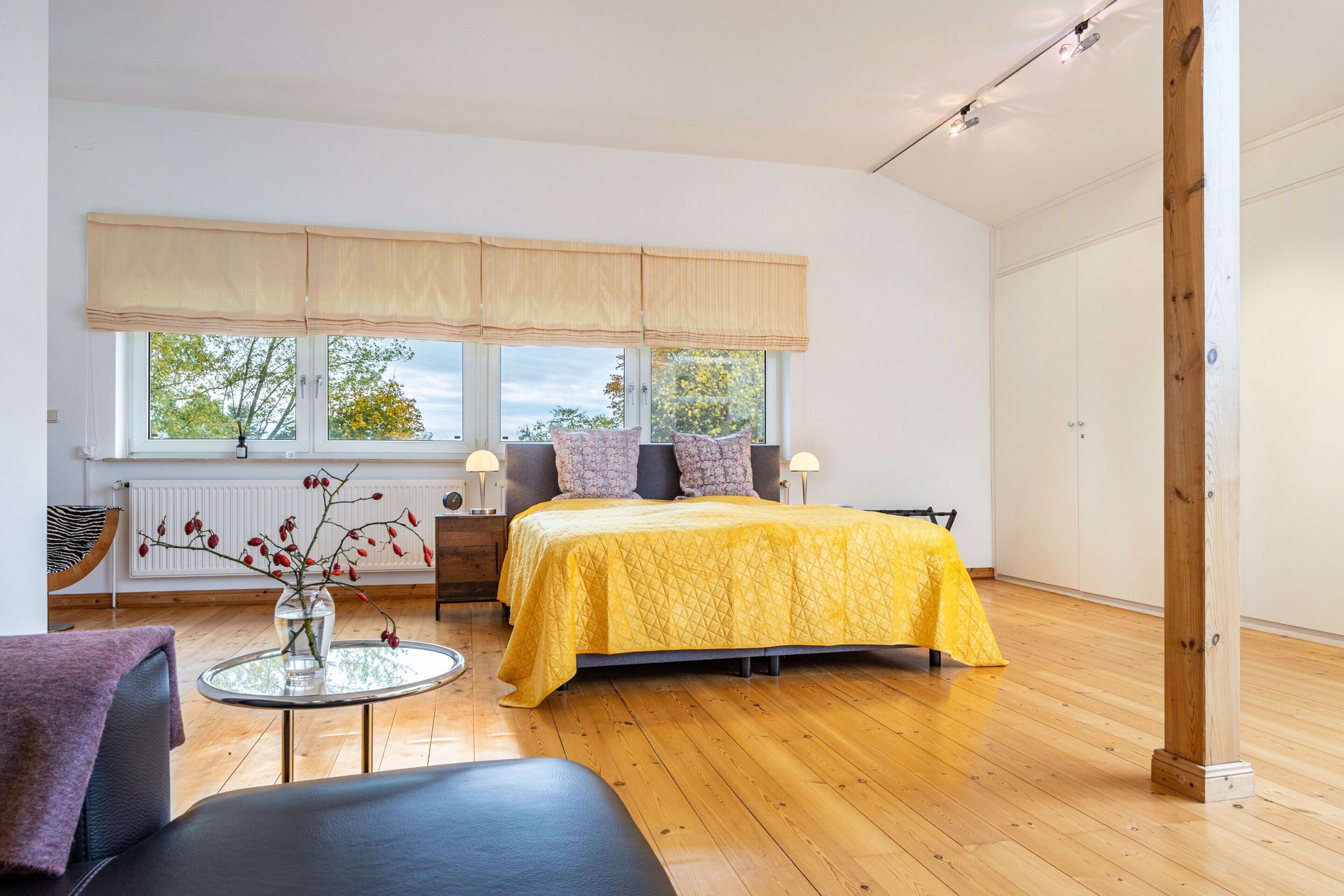 Quarto com cama, mesas de cabeceira, janela com cortinas, uma mesa redonda com um vaso e uma cadeira.