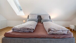 Een slaapkamer met een schuine plafond, dakraam, een bed met veel kussens en een nachtkastje met een lamp.