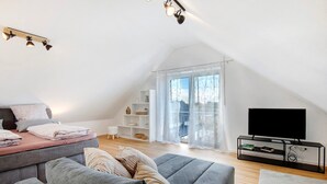 Een moderne loft slaapkamer met een schuine kap, een groot raam met lichte gordijnen, een flatscreen televisie op een zwart voetstuk, en een grijze bank met meerdere kussens.