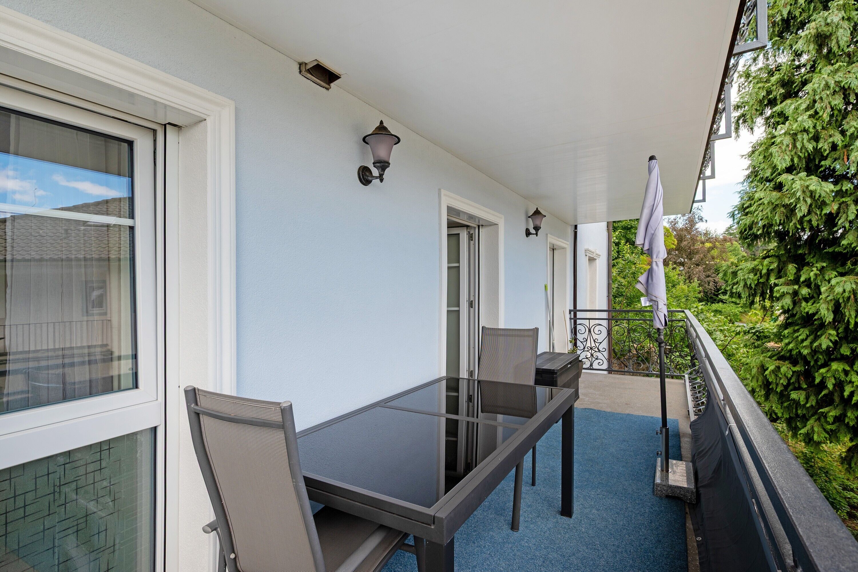 Een balkon met een glazen tafel en stoelen, een witte parasol en uitzicht op groen.