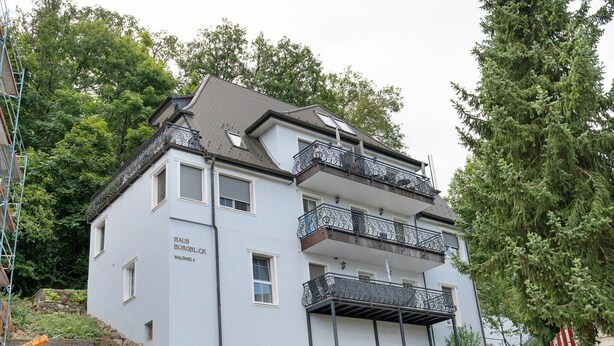 Un edificio de varios pisos con balcones, un muro de piedra y abundante vegetación circundante.
