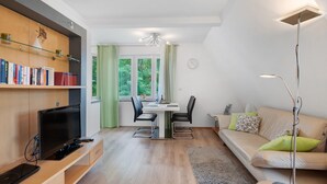 Una sala de estar moderna con un televisor de pantalla plana, un sofá con cojines, una zona de comedor con una mesa y sillas, y una estantería con libros.
