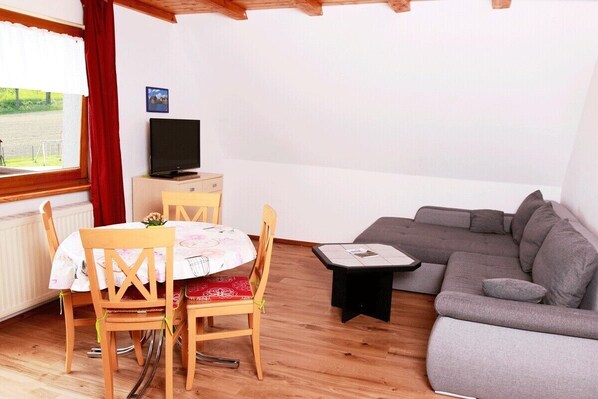 A room with a dining table, chairs, a sofa, a TV, and a window with a view of greenery.