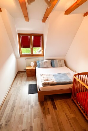 Un dormitorio con piso de madera, una cama con almohadas, una mesita de noche, un ventanal con persianas rojas y una cuna.