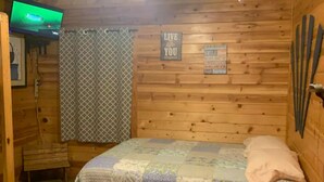 Une pièce chaleureuse aménagée dans une cabane en bois, avec un lit, un ventilateur de plafond, une télévision et des affiches inspirantes accrochées aux murs.