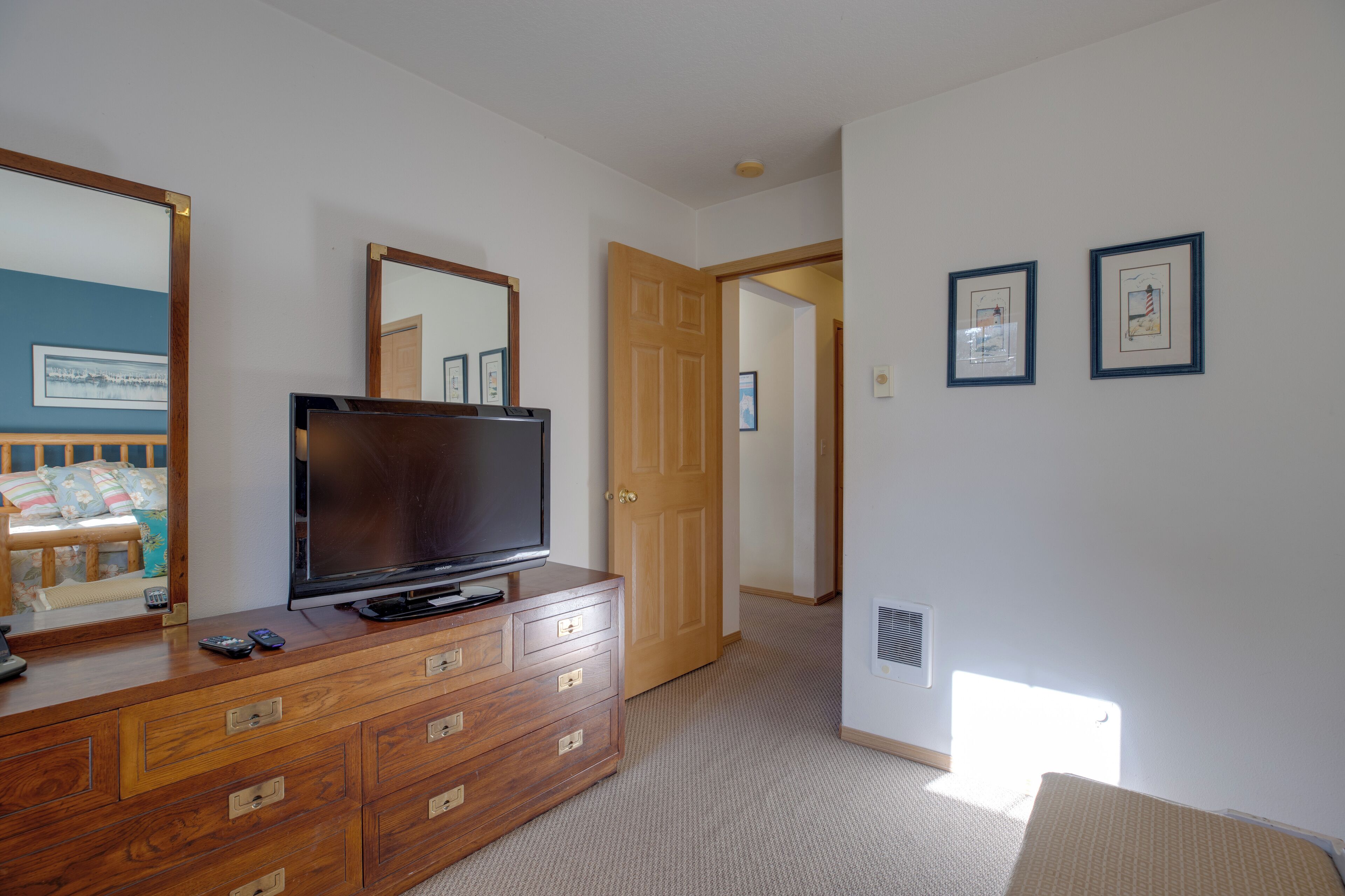 A bedroom with a wooden dresser, a television, and a large mirror. There is a door leading to another room and a small window.