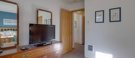 A bedroom with a wooden dresser, a television, and a large mirror. There is a door leading to another room and a small window.