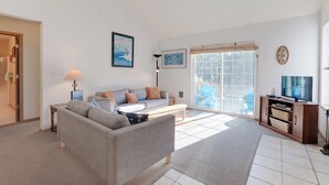 A living room with a sofa, coffee table, and TV stand. There is a window with blinds and a door leading to another room. - Kiwanda Cabana - near beach, cute house! Dog OK. (Pacific City)