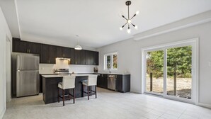 A modern kitchen with dark cabinets, a stainless steel refrigerator, a central island with bar stools, and a large window offering a view of the outdoors. - Amazing stay near Hamilton-Brantford- Beach Port Dover (Norfolk)